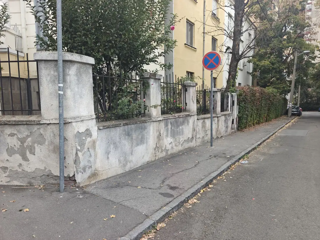 Pogled niz trotoar na staru zidanu i metalnu ogradu pre rušenja